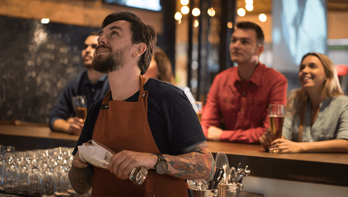 Allente En bartender og en gruppe mennesker i en bar ser på en TV-skjerm